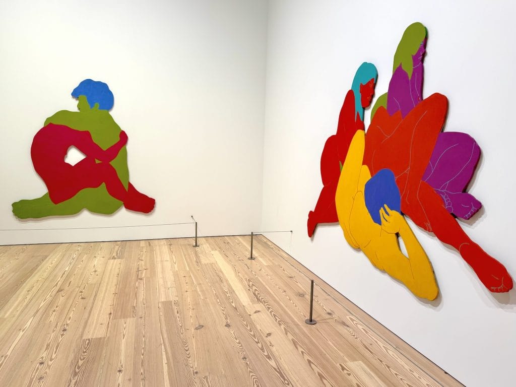 An installation view of Carmen de Monteflores’s work at the Whitney Biennial, featuring large, brightly colored cut-out figures in various poses mounted on two adjacent gallery walls.