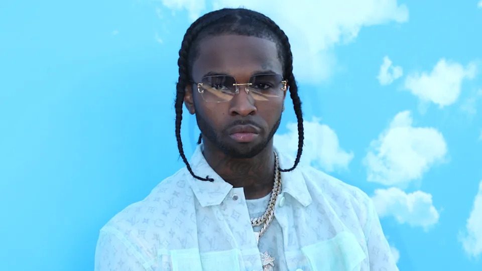 Pop Smoke with braids and sunglasses stands against a blue sky background with clouds, wearing a patterned shirt and necklaces.