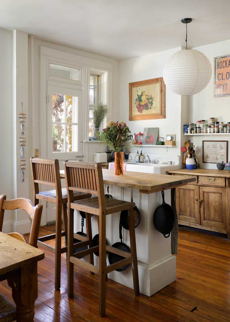 Cozy kitchen with wooden bar stools, a rustic table, potted flowers, and hanging cookware near a window.
