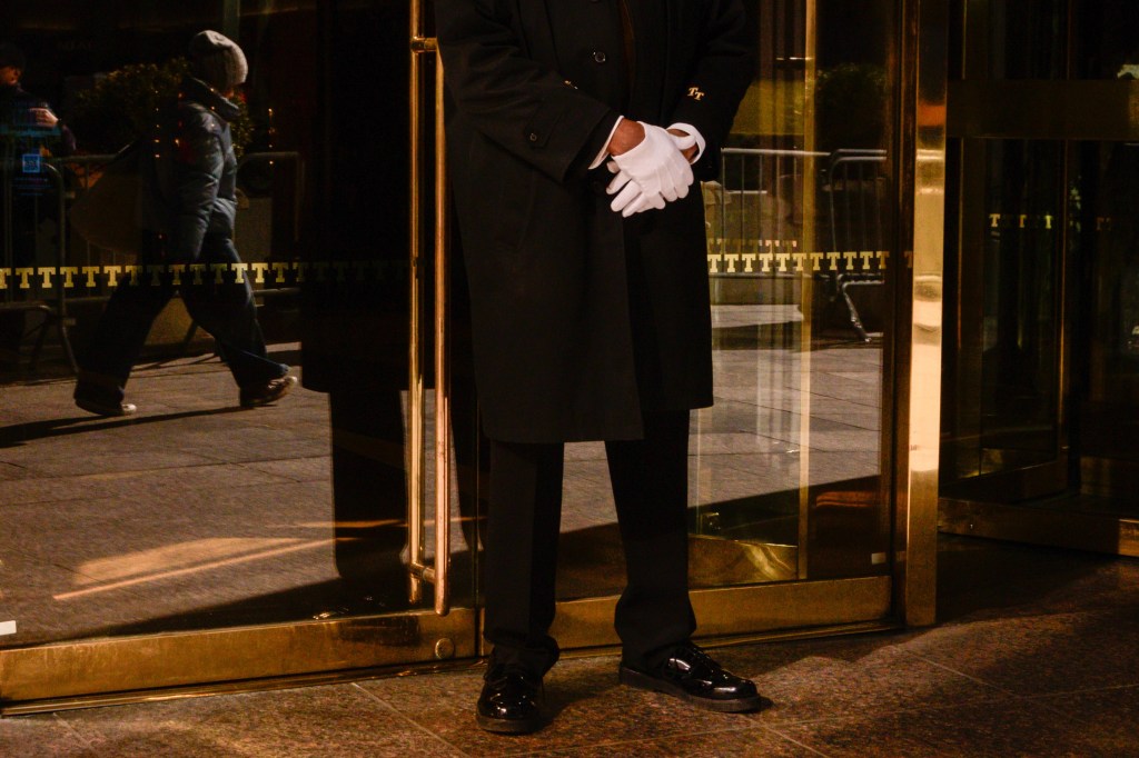 A doorman in a black coat and white gloves stands outside of Trump Tower.