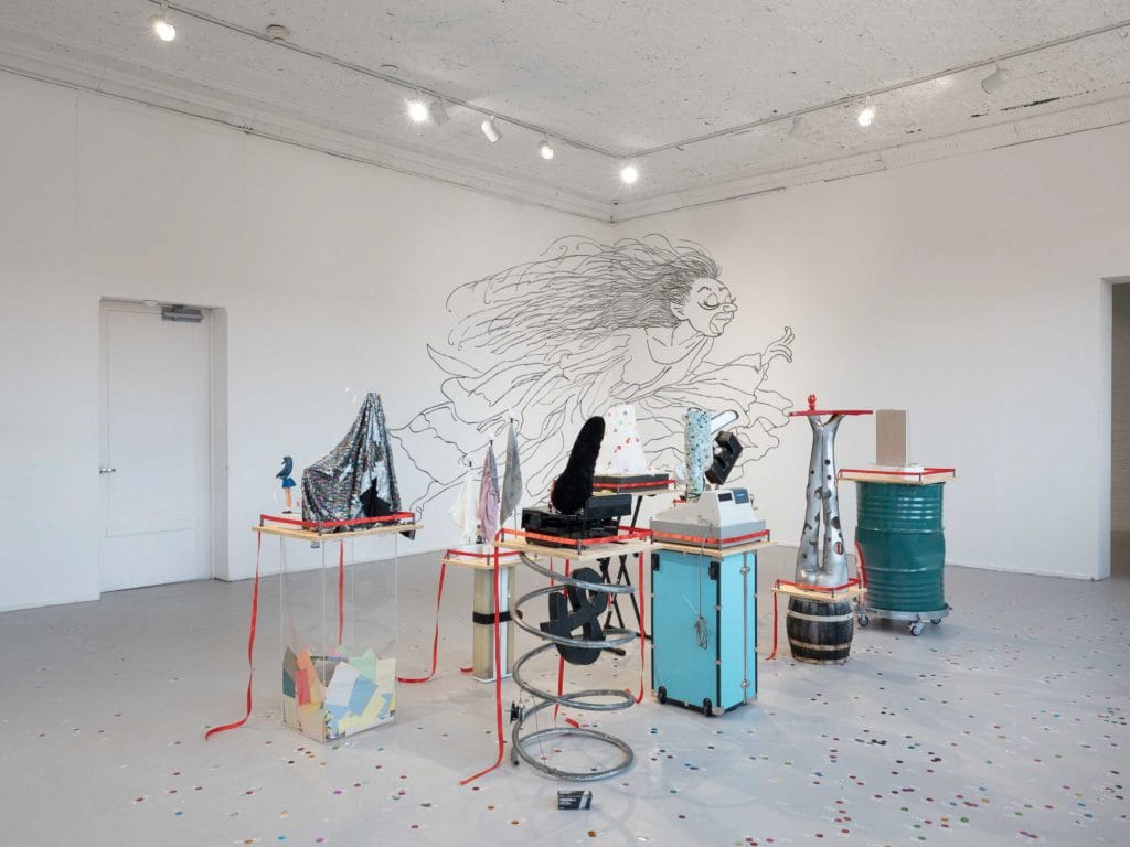 A photograph of six standing scuptures of colleaged items in a white walled gallery with a line drawing of a running woman spreading over the corner behind them