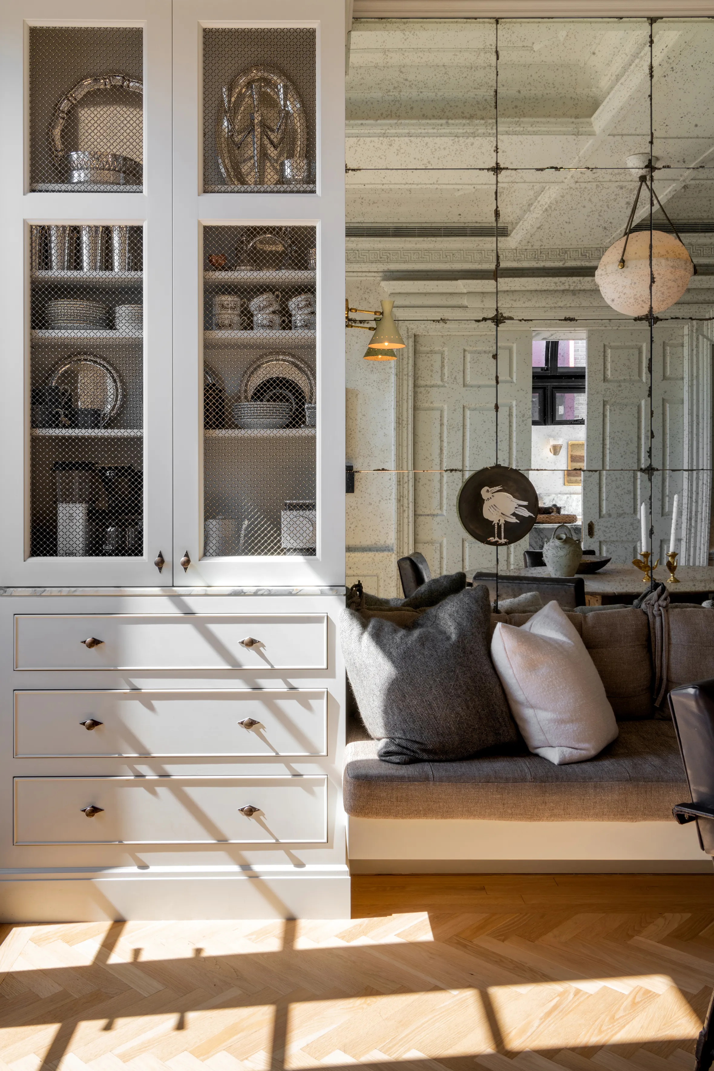 Built-in white cabinets with wire mesh doors next to a built-in bench with throw pillows, in a room with an antiqued mirror wall.