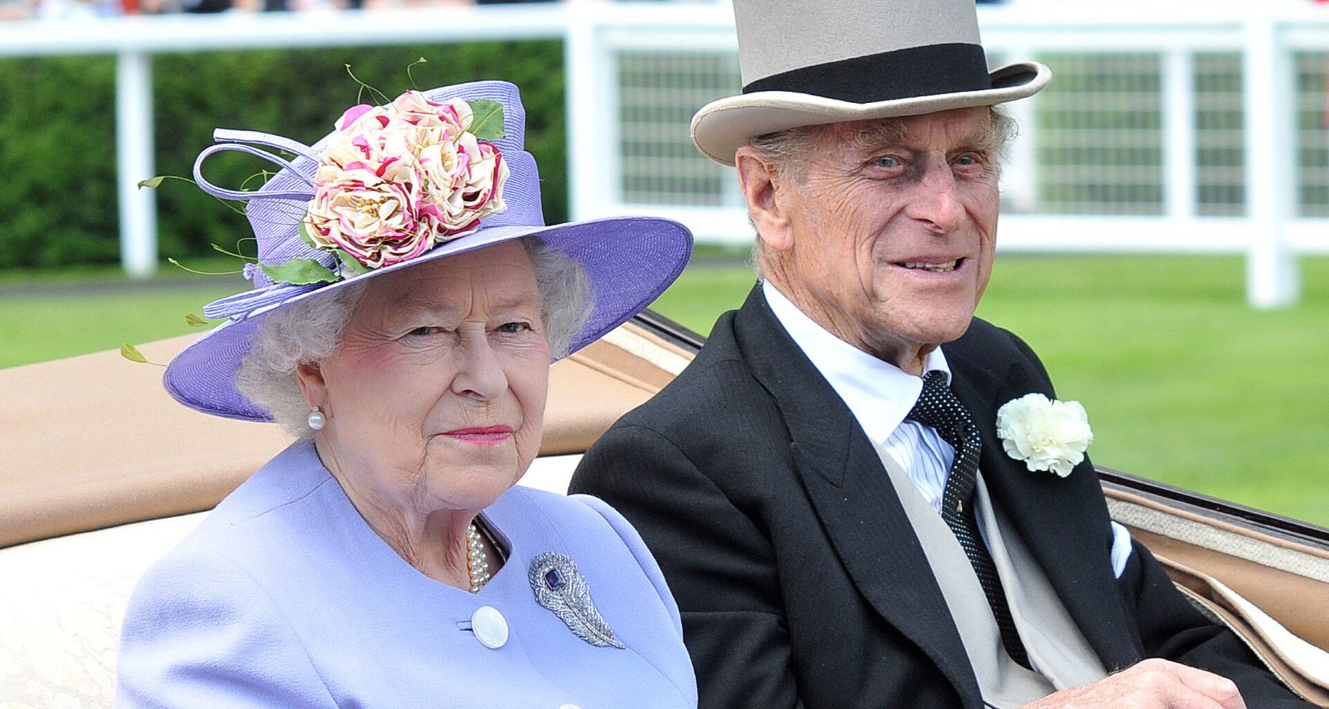 Queen Elizabeth wearing a lilac hat and coat and sitting next to Prince Philip who wears a top hat