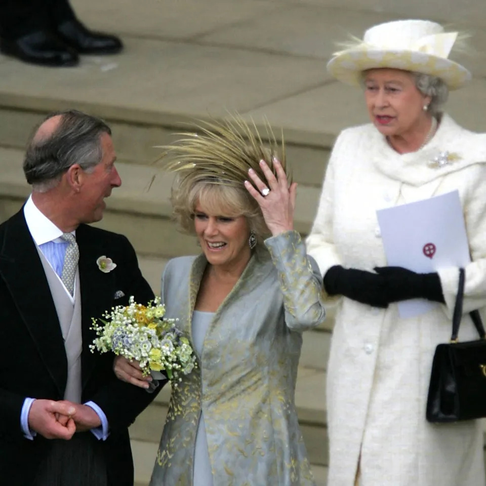 King Charles linking arms with Queen Camilla, holding on to her hat on her wedding day, with Queen Elizabeth standing behind her.