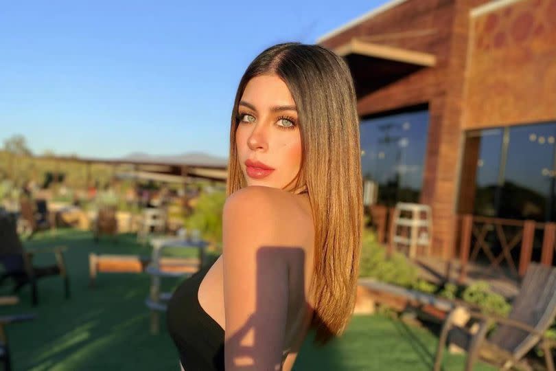 An individual with long hair is standing outdoors, holding a camera. The setting appears to be a garden or patio area, featuring various plants and a building in the background. The scene suggests a leisurely or relaxed atmosphere with daylight.