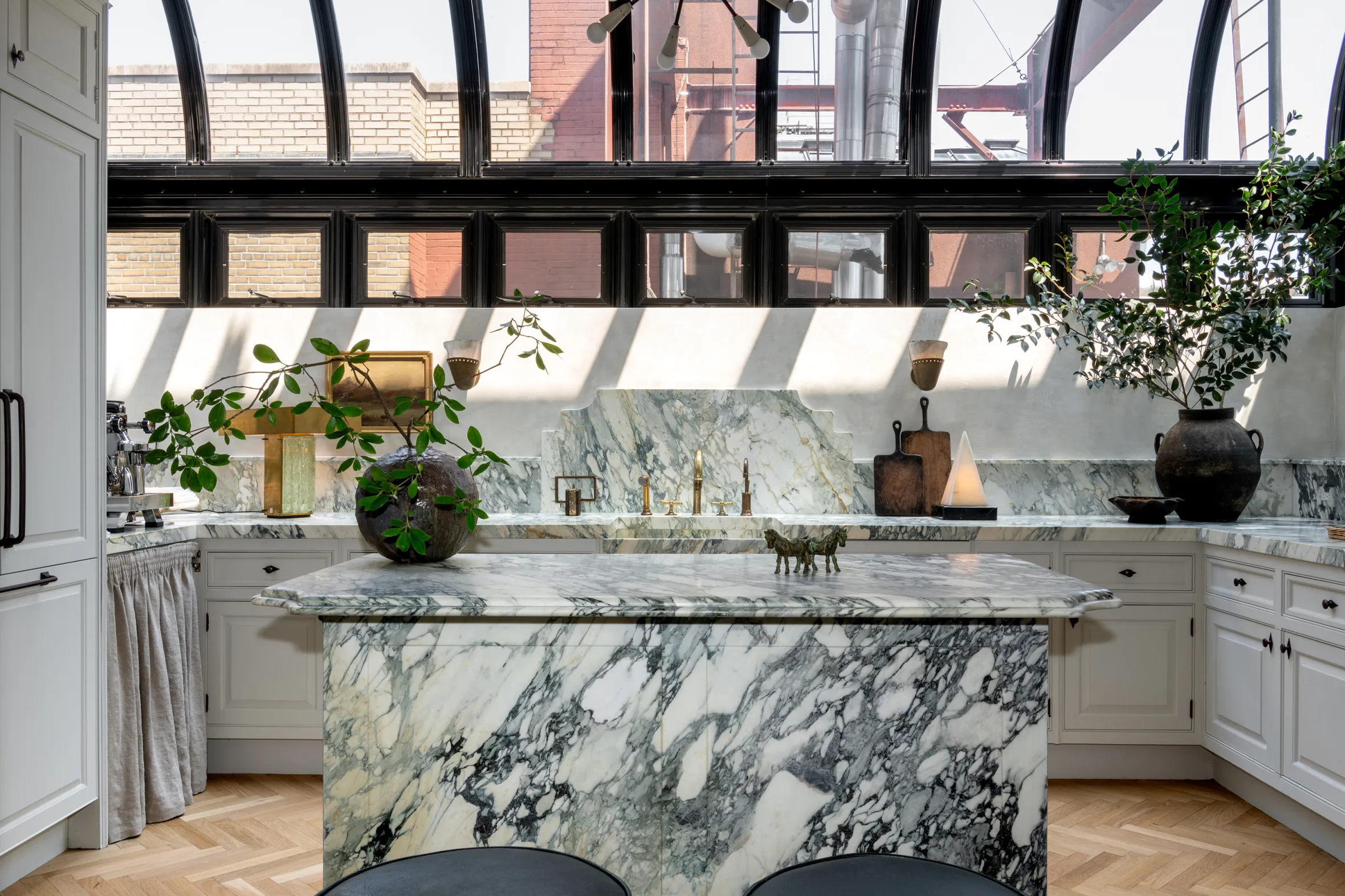 A kitchen with white cabinets, marble countertops and backsplash, a large marble island, and large windows that offer a view of red brick buildings.