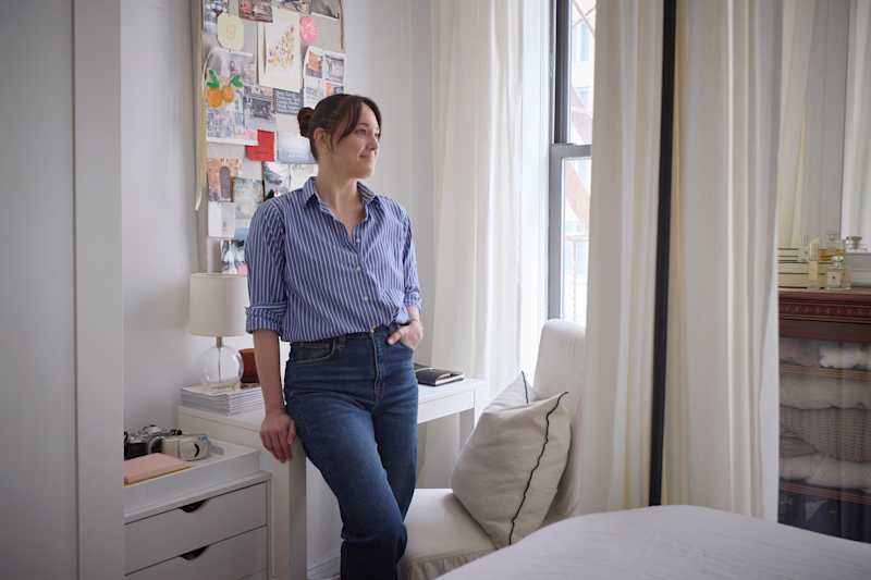 A woman in a blue striped shirt leans against a white desk in a bright room, with a mood board and window behind her.