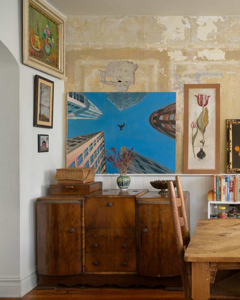 Wooden sideboard with a basket, surrounded by various framed artworks and a dining table with a chair.