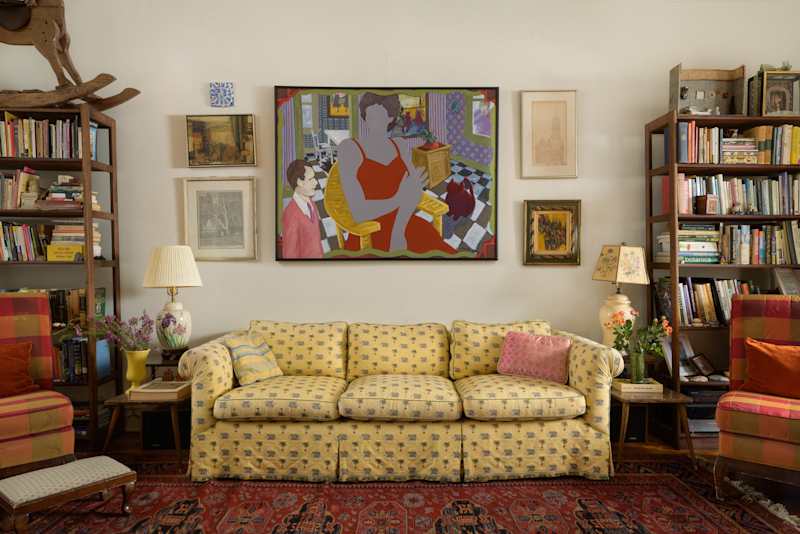 Cozy living room with a yellow patterned sofa, colorful artwork, bookshelves, and a floral lamp.