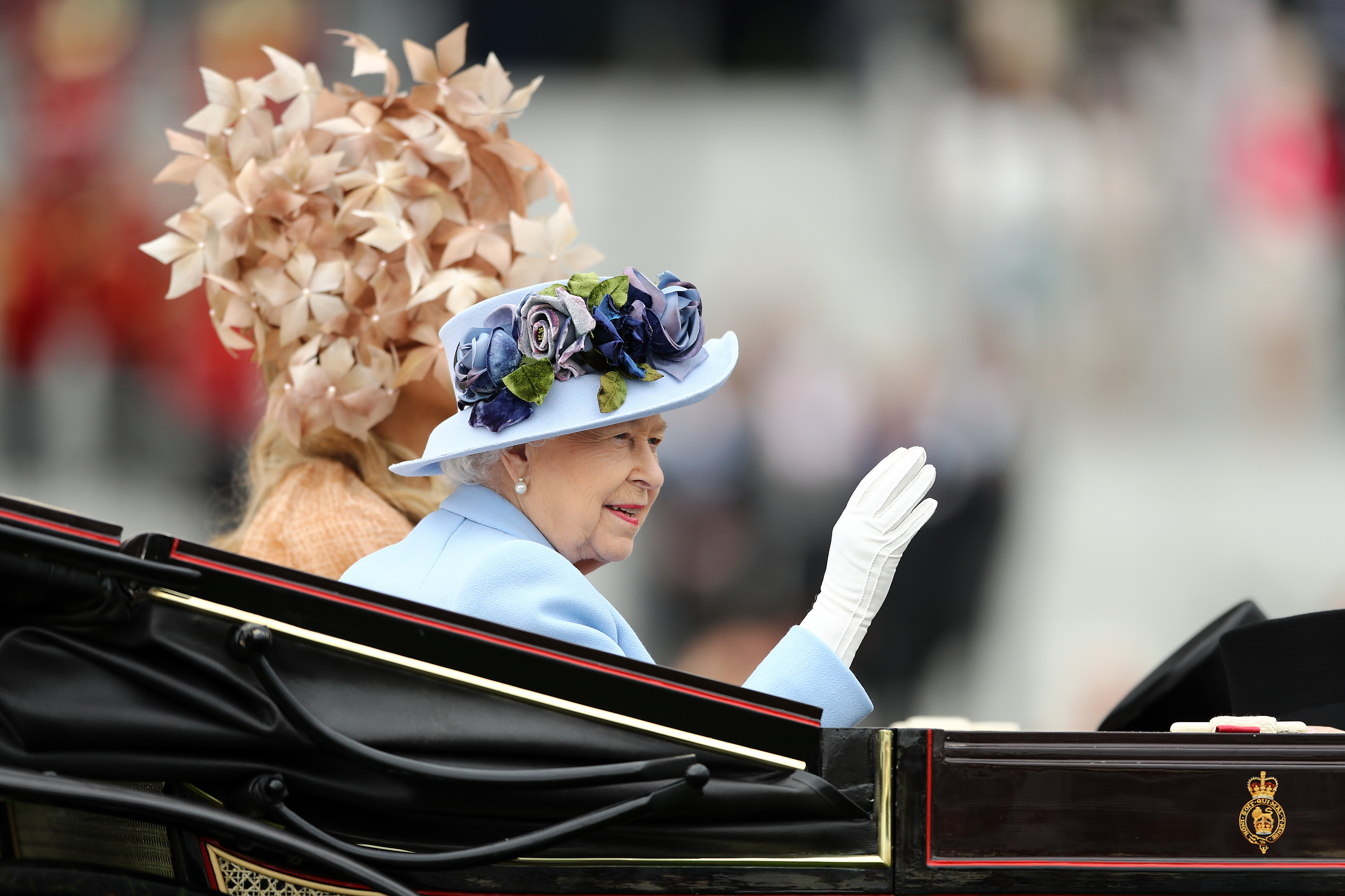 Queen Elizabeth wearing a blue hat and waving in a carriage