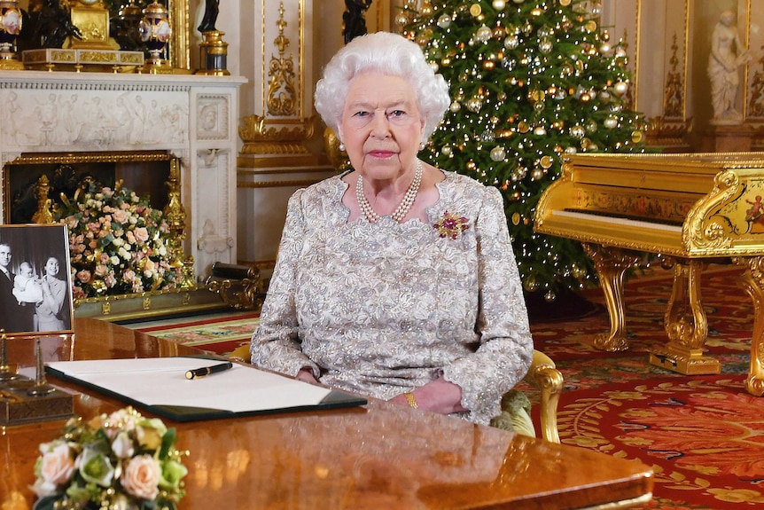 Queen Elizabeth II sits at a desk in  a grand drawing room with a Christmas tree behind her