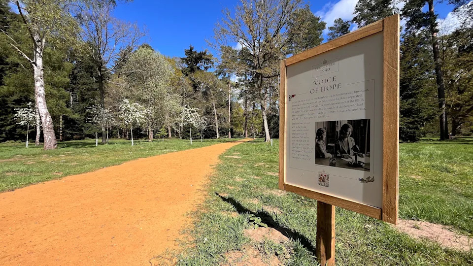 A wooden sign is on the right and it has a black and white picture and text. There is a path on the left which leads to a woodland area.