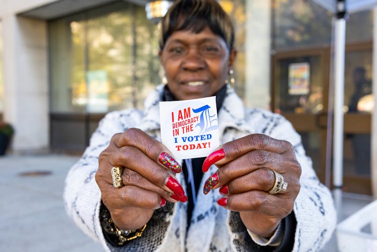 A Black woman with a great red manicure holds a sticker that reads 'I am democracy in the D. I voted today.'