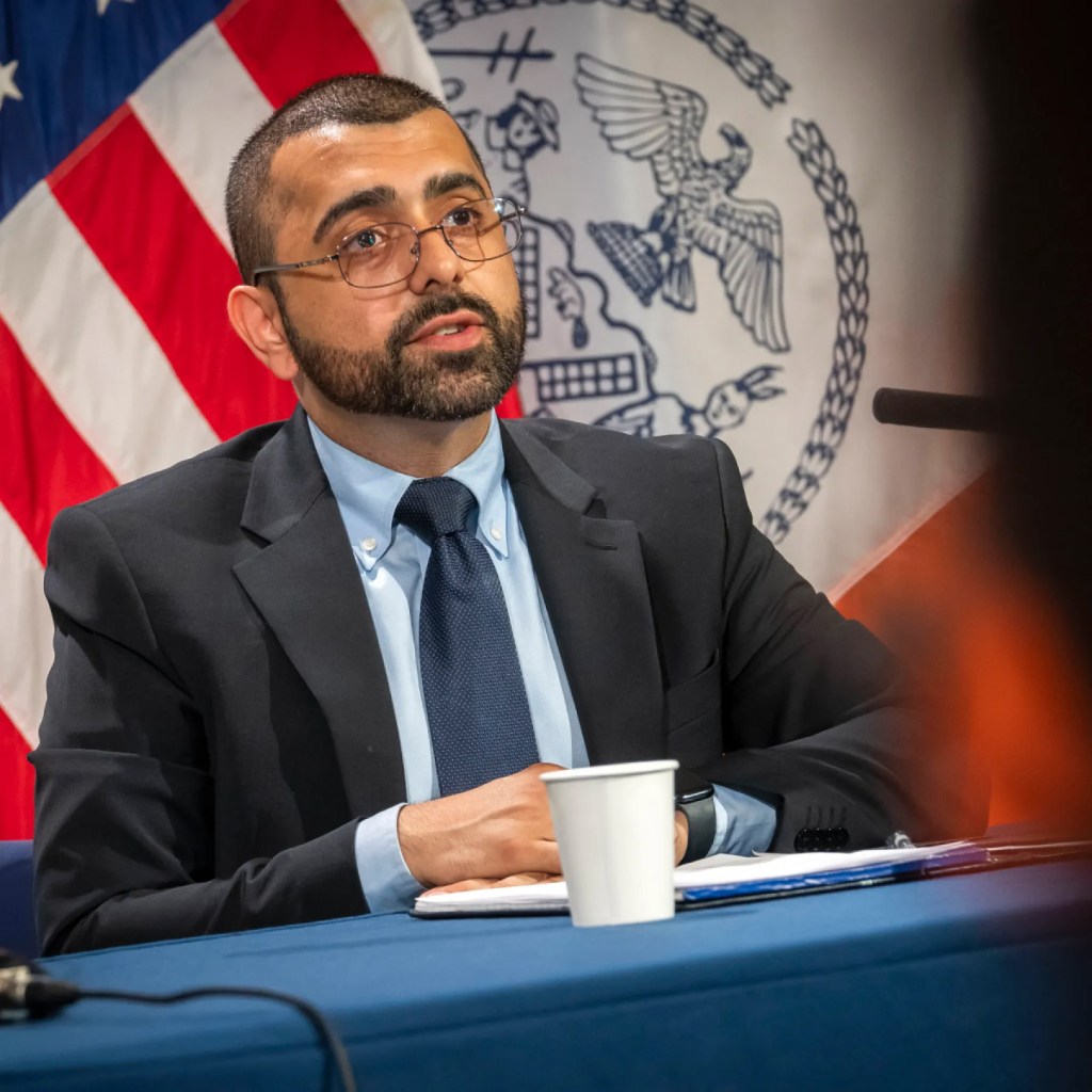 Hassan Naveed, former director of NYC's Office for the Prevention of Hate Crimes, sitting at a table with an American flag and the NYC seal in the background.