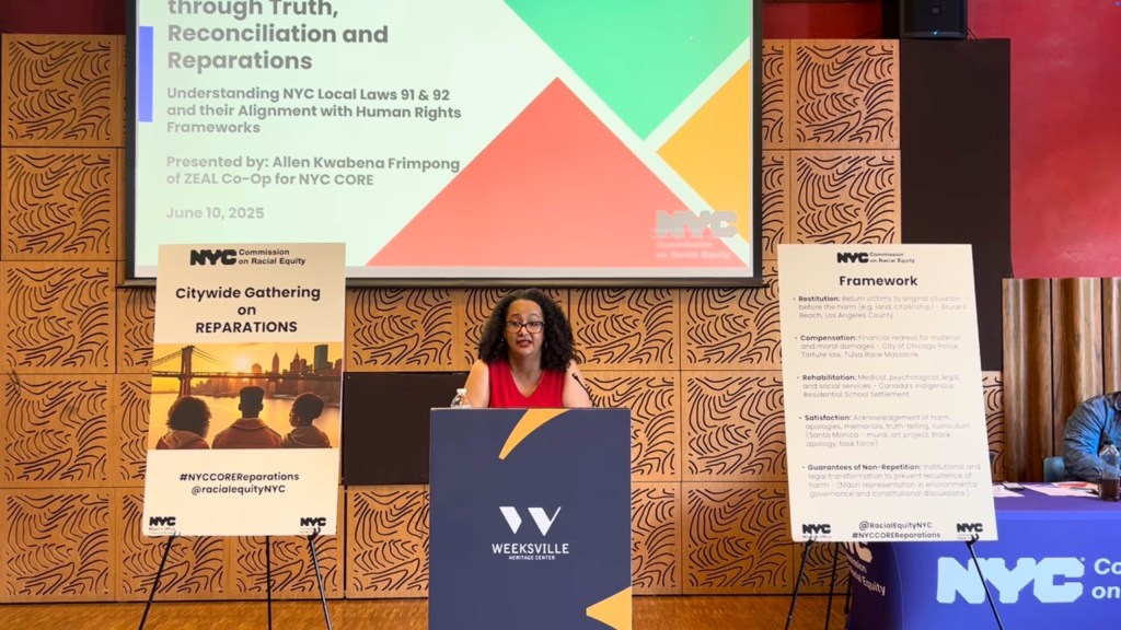 A woman speaks at a podium in front of a presentation screen and two informational signs for the NYC Commission on Racial Equity's citywide gathering on reparations.