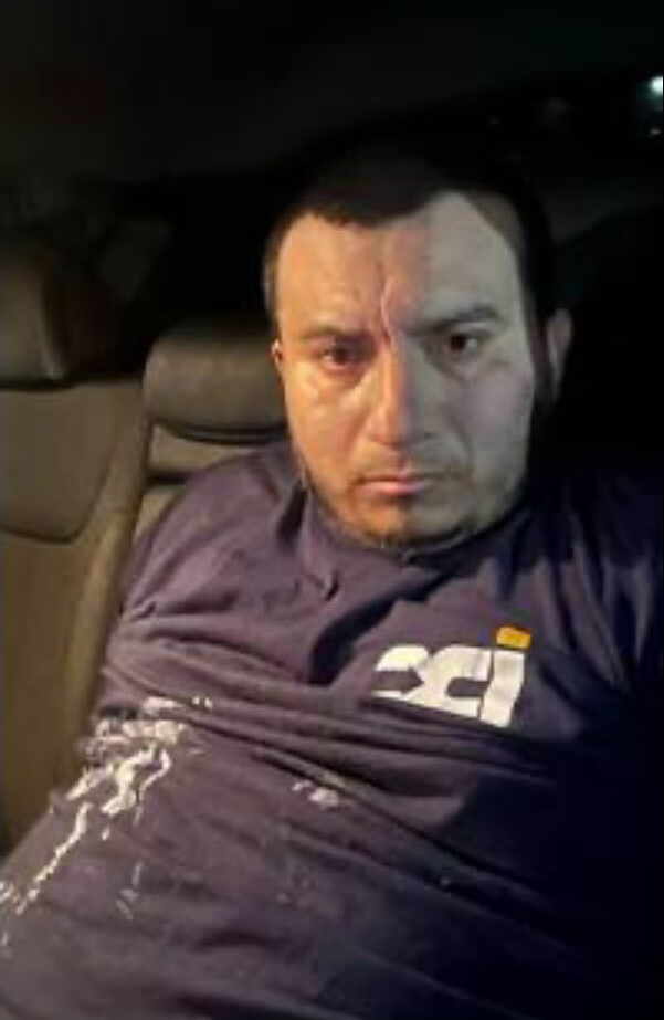 Man with a serious expression, wearing a navy blue shirt with a white and orange logo, sitting in a vehicle.