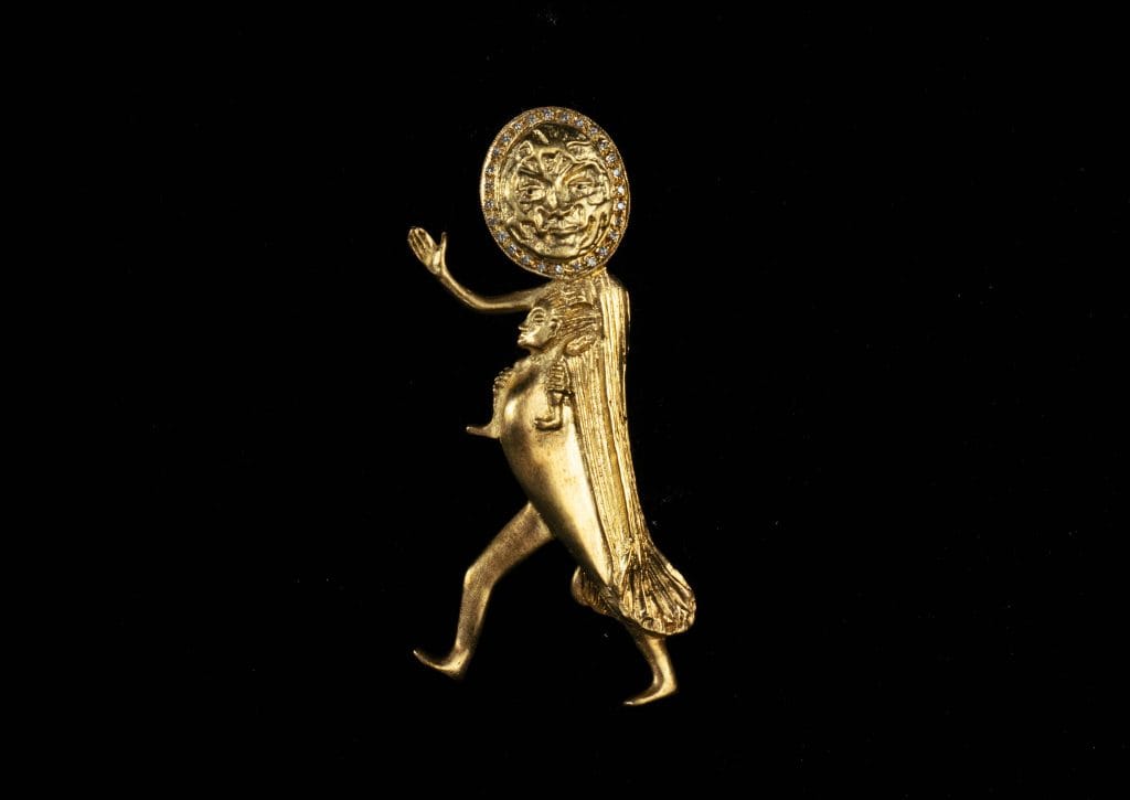A small, gold-toned sculptural figure with a round, sun-like face framed by a halo. The figure has a stylized, elongated body with one arm raised in a gesture and one leg lifted as if walking. A smaller human-like form appears integrated into its torso. The sculpture is set against a solid black background.