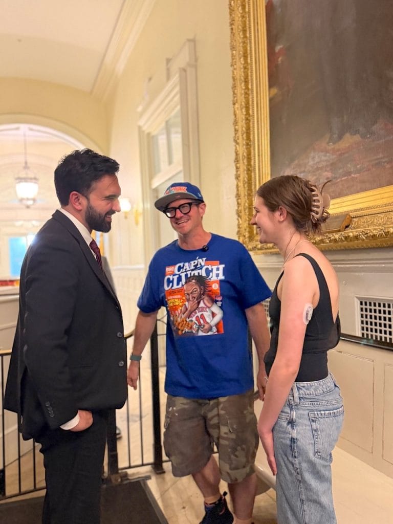 Three people stand talking in a grand, high-ceilinged hallway next to a large gold-framed painting. A man in a suit faces a man in a graphic T-shirt and a woman in jeans.