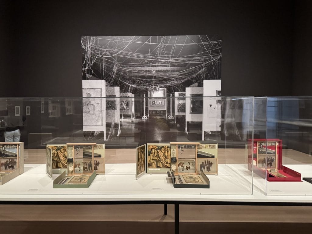 Museum display of Marcel Duchamp’s Boîte-en-valise sets in glass cases, positioned before a large black-and-white photograph of the 1942 
