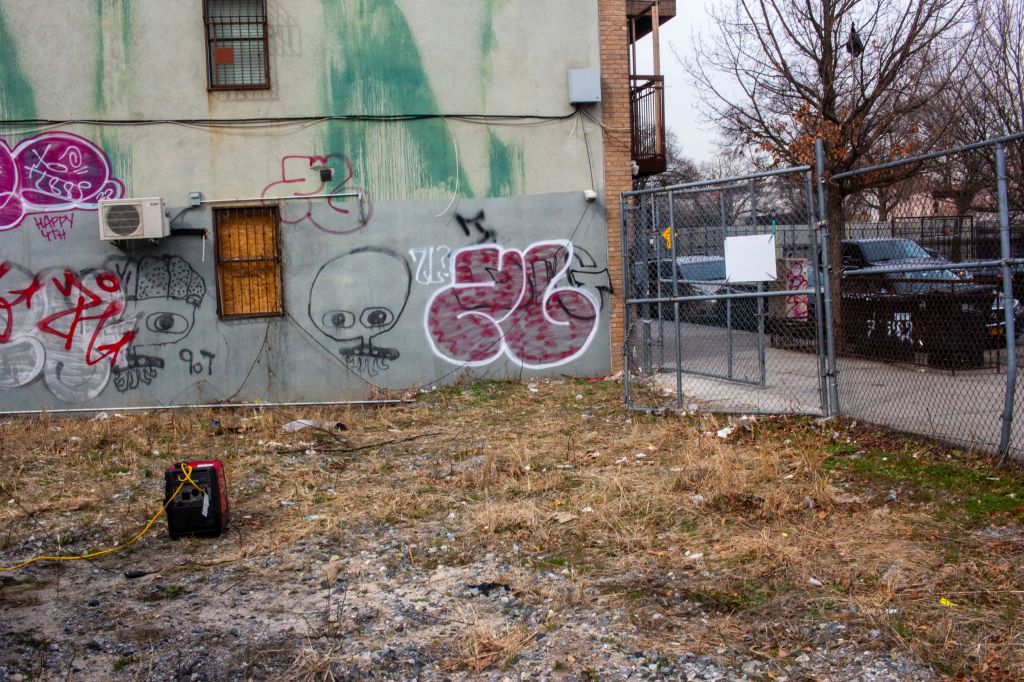 Vacant lot in Brooklyn with a graffiti-covered building, a chain-link fence, and a generator.