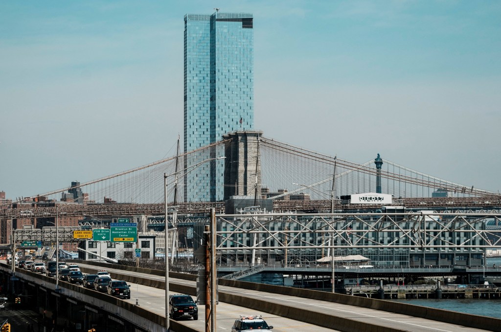 A motorcade of black SUVs and police cars travels on a multi-lane highway, with the Brooklyn Bridge and a tall glass skyscraper in the background.