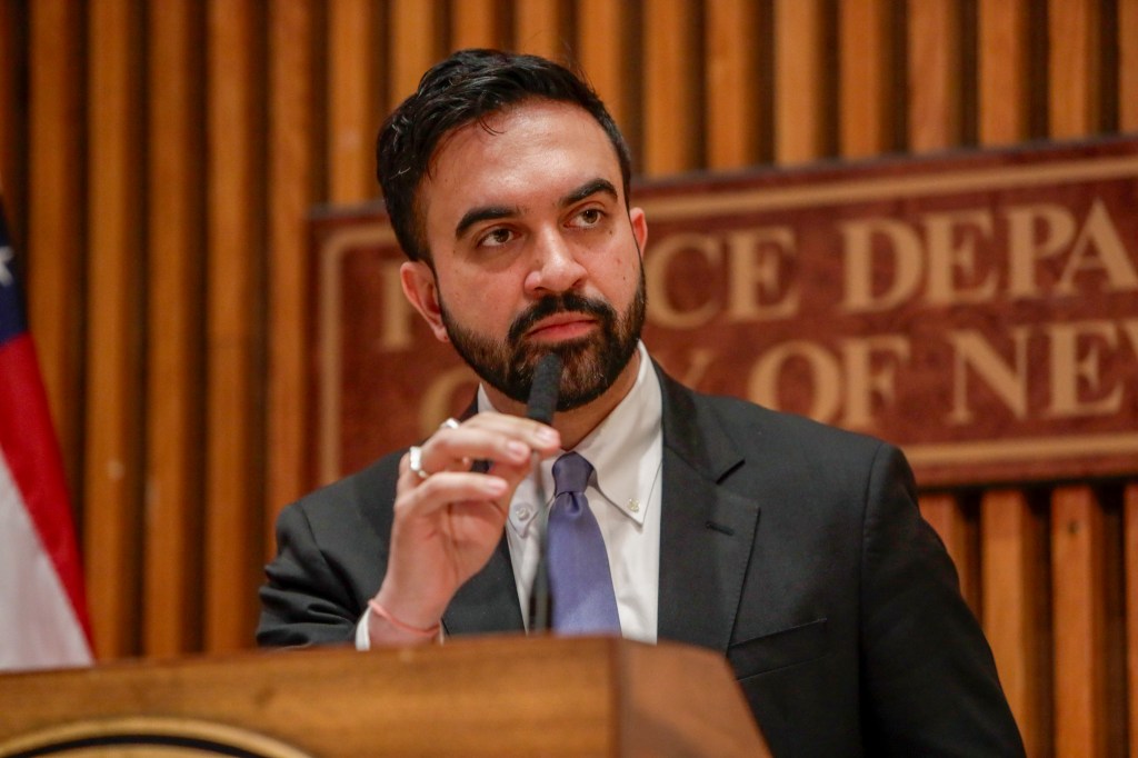 Zohran Mamdani speaking into a microphone at a press conference.