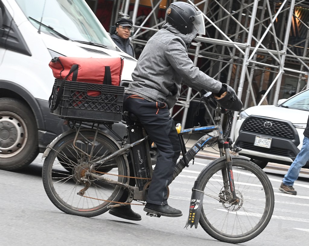 A food delivery worker on an e-bike, wearing a helmet, jacket, and dark pants, rides past a white van.