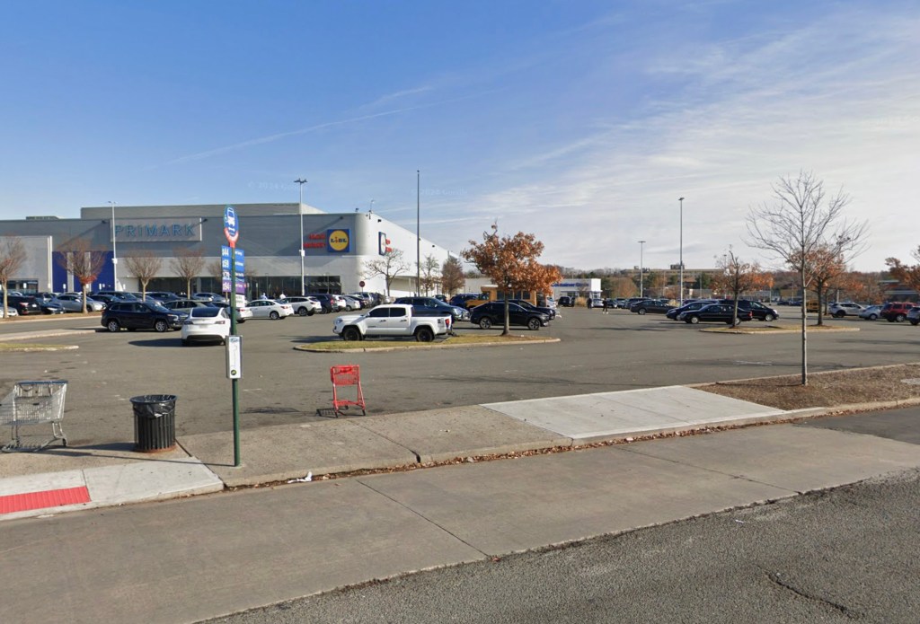A bus stop sign is visible on a sidewalk bordering a large parking lot in front of a Primark and Lidl store.