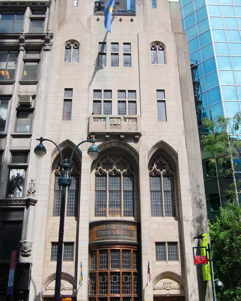 The facade of the Swedish Church of New York, a light-colored stone building with many windows, next to a modern glass building.