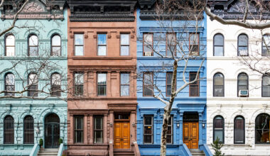 Four colorful Brooklyn condo brownstone townhouses in a row, painted green, brown, blue, and white, with bare trees in front and classic architectural details, each featuring stoops and wooden front doors.