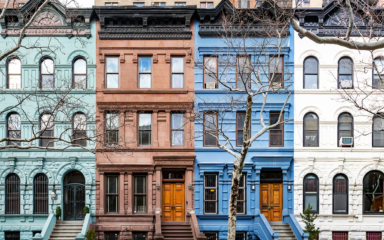 Four colorful Brooklyn condo brownstone townhouses in a row, painted green, brown, blue, and white, with bare trees in front and classic architectural details, each featuring stoops and wooden front doors.