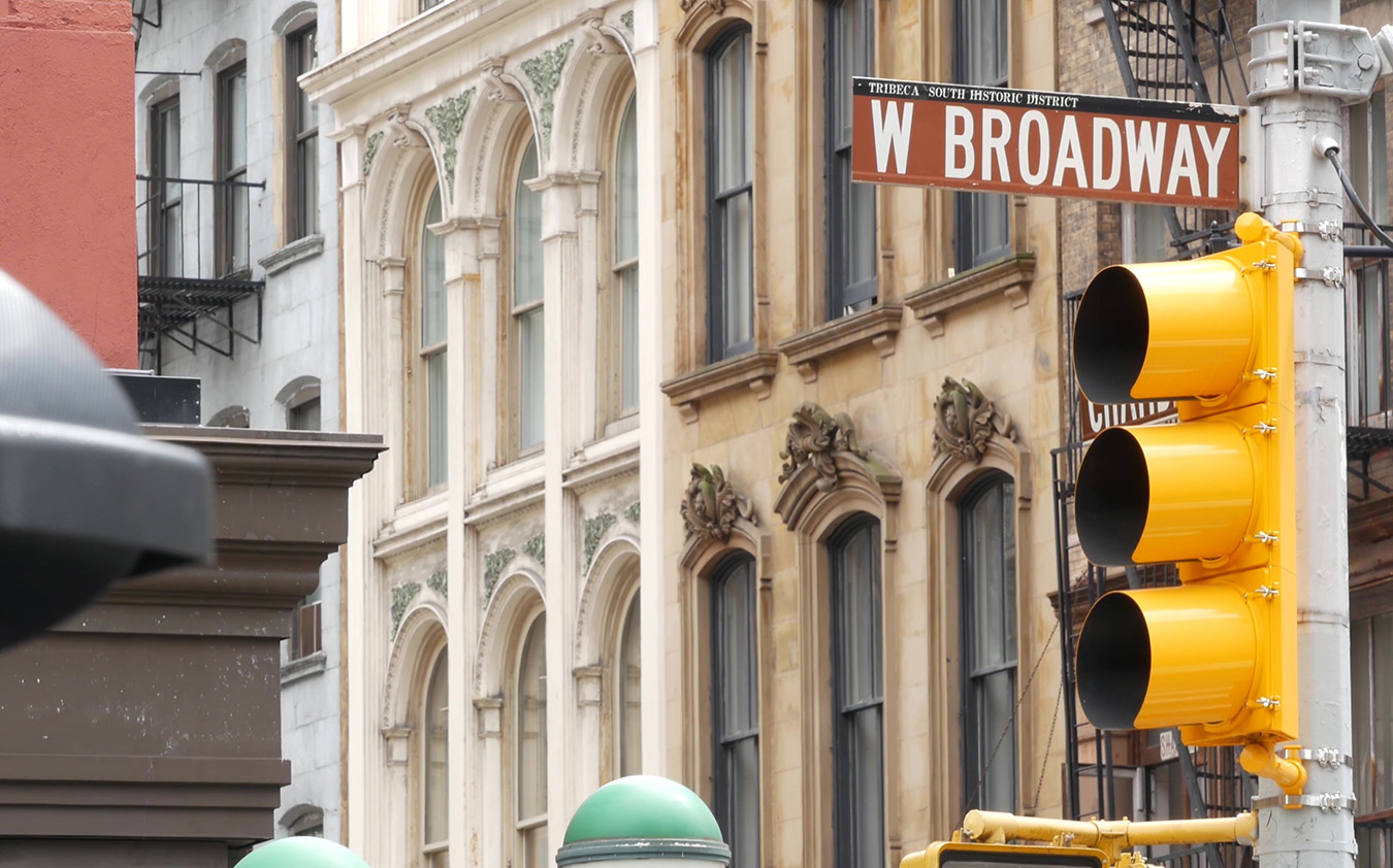 A close-up of a street sign reading W Broadway and a yellow traffic light, set against historic stone buildings with arched windows—capturing the charm of the Manhattan real estate market in an urban setting.