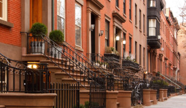 A row of classic brownstone townhouses with black iron railings and neatly trimmed shrubs lines a quiet city street, bathed in warm daylight.