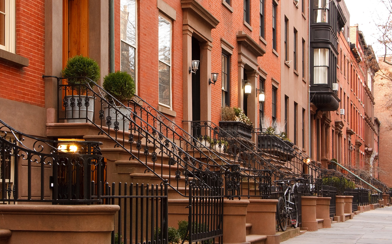 A row of classic brownstone townhouses with black iron railings and neatly trimmed shrubs lines a quiet city street, bathed in warm daylight.