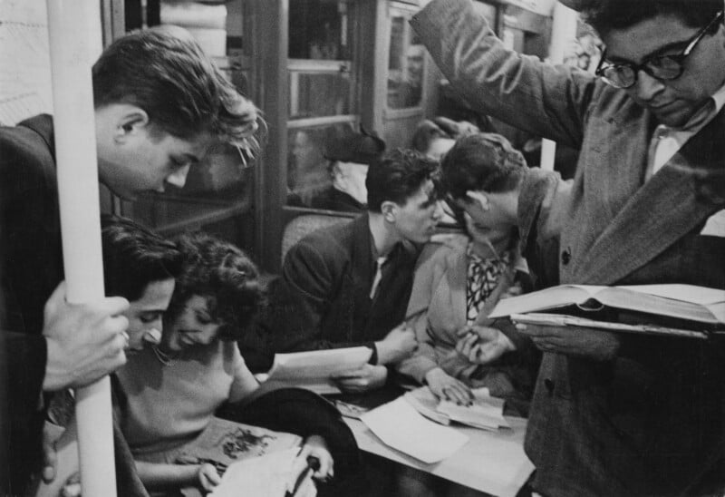 A group of young people, some standing and some sitting, read books and papers on a crowded subway train. The scene captures focused study and quiet interaction among the passengers.