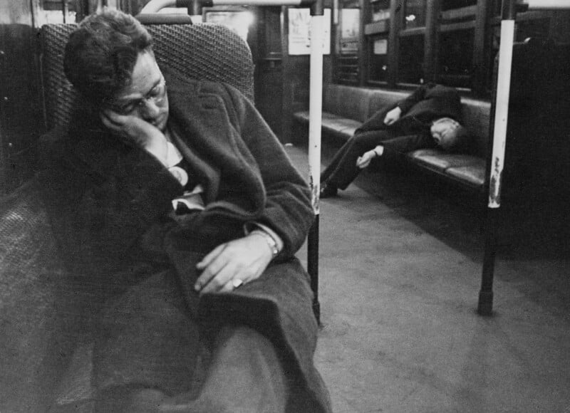 Two people sleep on an empty subway train; one slumps on a seat in the foreground with their head resting on their hand, while the other reclines on a bench in the background. The scene appears quiet and dimly lit.