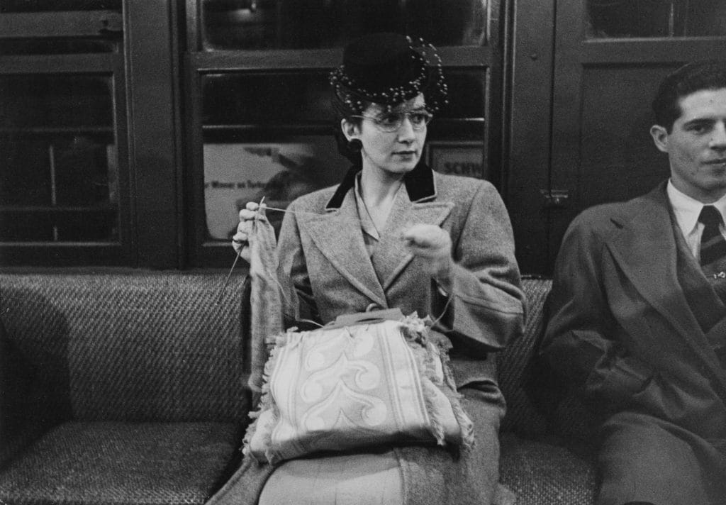 A black and white Stanley Kubrick photo depicting a fashionably dressed woman sitting on a subway train with a a textile project she's working on in her lap