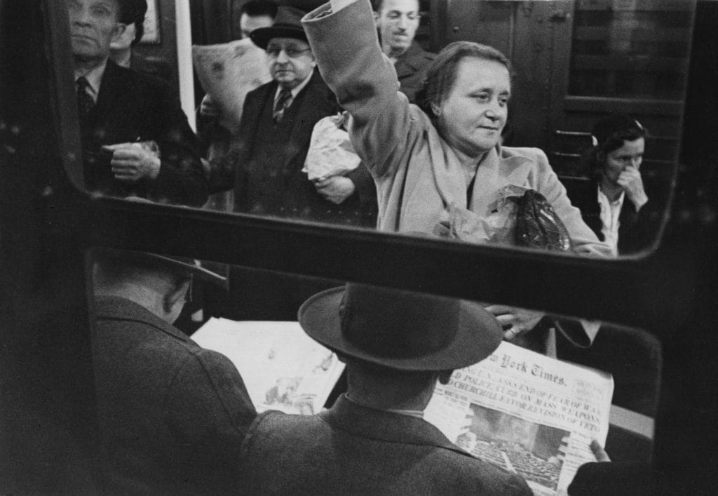 A black and white photograph depicting a crowded subway train with a woman standing, holding on to a strap, as seen through the train window