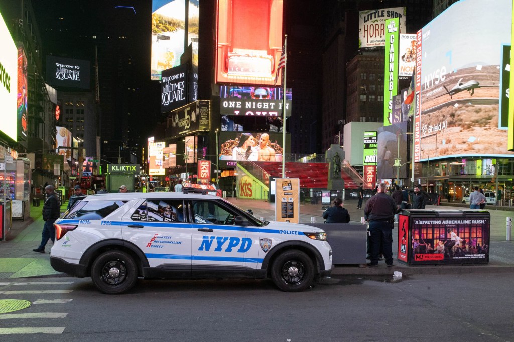 An NYPD police car parked at night in Times Square, New York City, surrounded by large, brightly lit billboards and advertisements.
