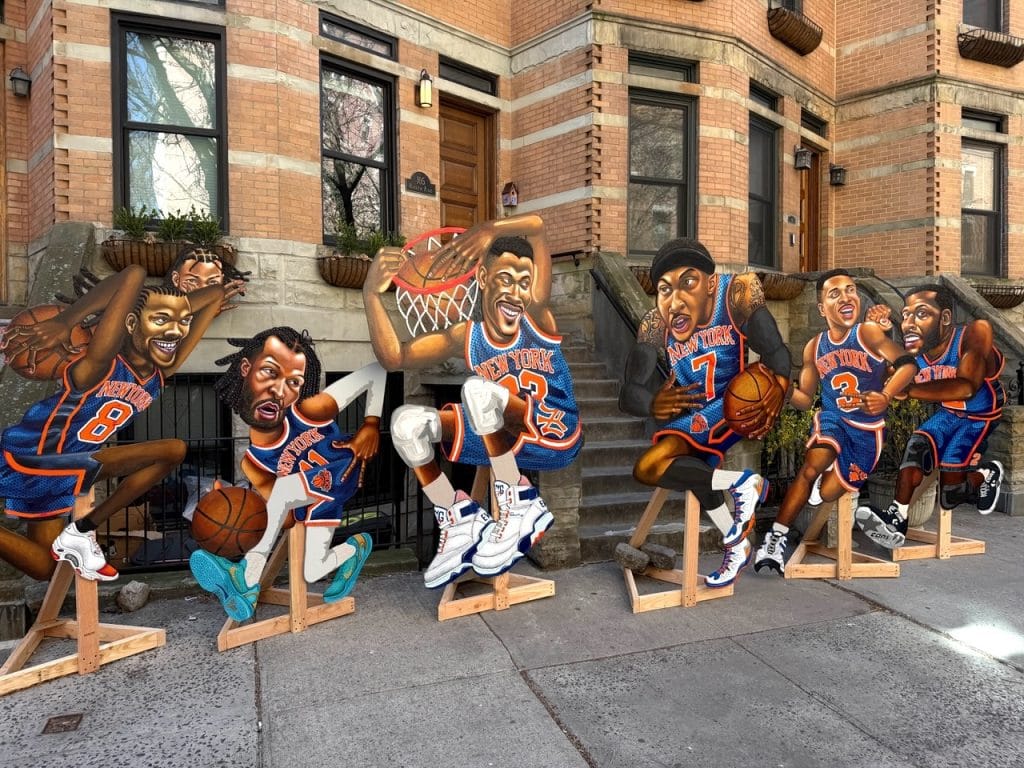 Six hand-painted wooden cutouts of legendary New York Knicks players, rendered in a colorful caricature style, are displayed on a city sidewalk in front of brownstone buildings.