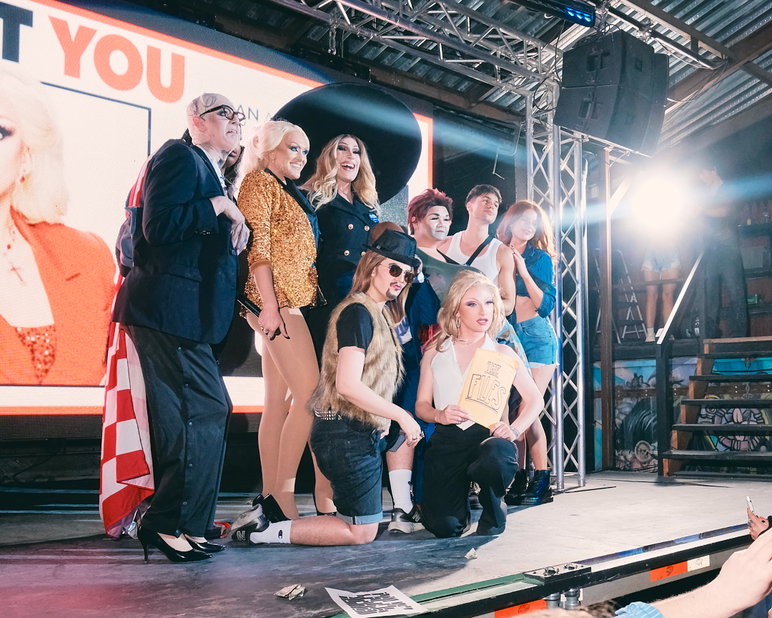A group of drag performer post onstage in front of a large projection screen showing a promotional image for the event.