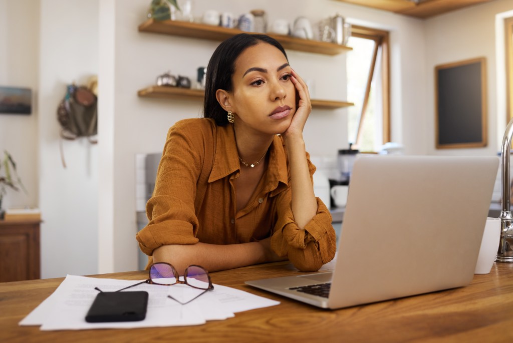 A tired woman rests her head in her hand while looking at her laptop.