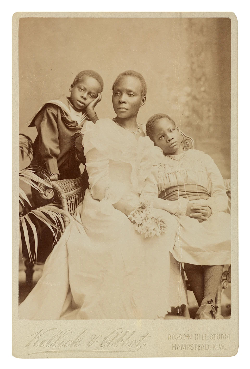 Sepia-toned vintage photo of three individuals in ornate period clothing seated on wicker furniture with floral and leafy decor