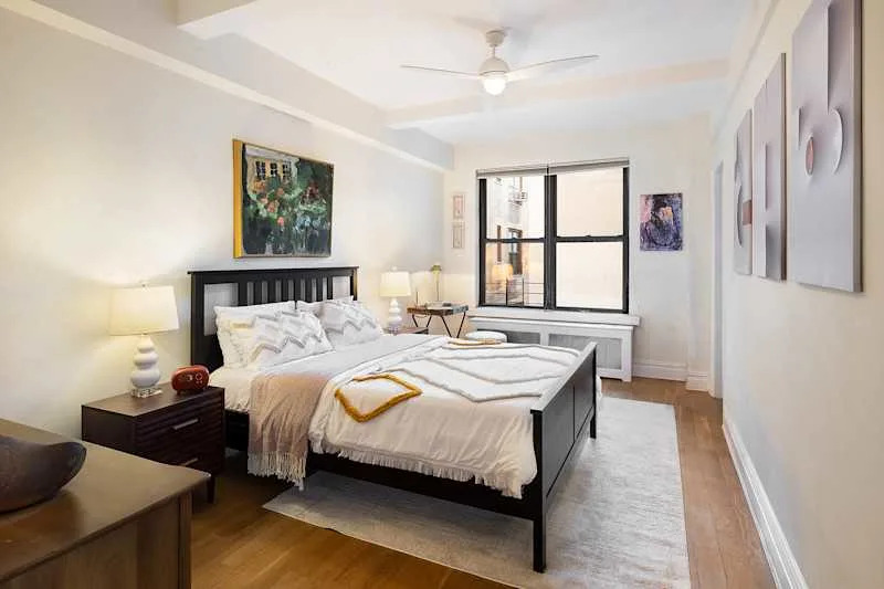 Bright bedroom featuring a black wooden bed with white bedding, a colorful painting, and large windows.