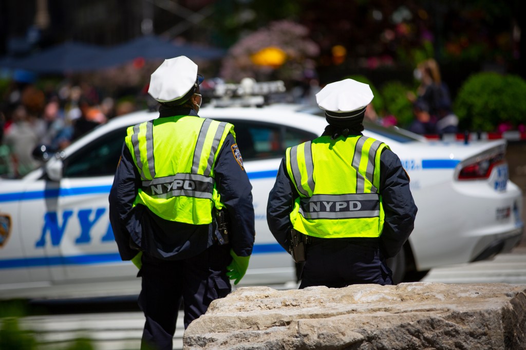 Two NYPD officers in reflective vests standing on a New York City street with a police car in the background.