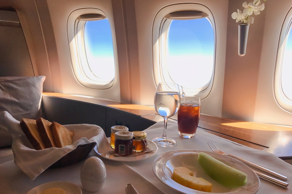 First-class dining on a Cathay Pacific flight with toast, fruit, beverages, and jams, beside three airplane windows.
