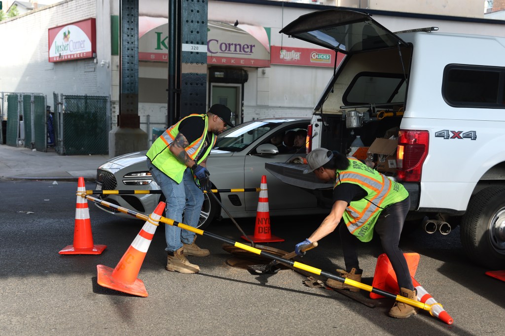 Two workers in safety vests are using tools to lift a manhole cover on a city street, with a white truck and parked car nearby.