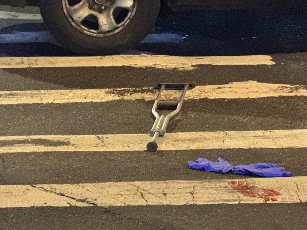 A single crutch, purple gloves, and bloodstains on a crosswalk, with a car tire in the background, indicating a hit-and-run incident.