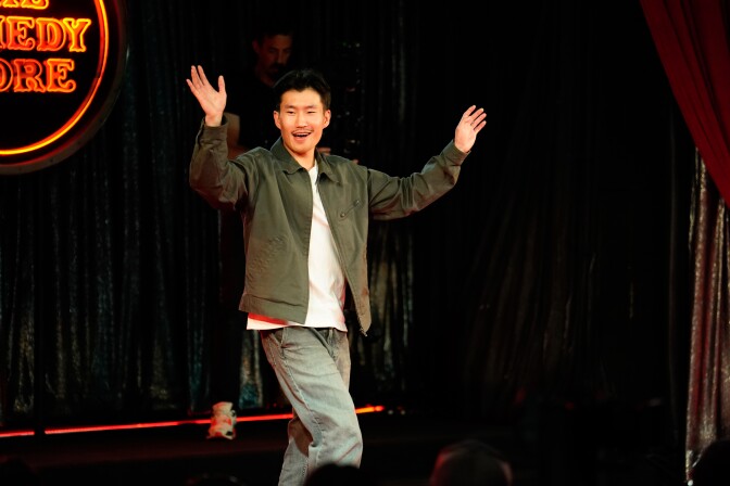 An Asian man with black hair, wearing a white t-shirt, jeans and army green jacket walks onto a stage smiling and with his hands up. Behind him are black curtains and a red/orange neon sign that partially reads "The Comedy Store."