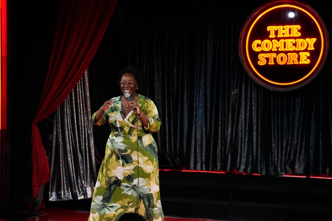 A Black woman on stage holding a microphone and smiling. She's wearing glasses and a long green and white floral dress. Behind her are black curtains and an orange neon sign that reads "The Comedy Store." A red velvet curtain is pulled back to her right.